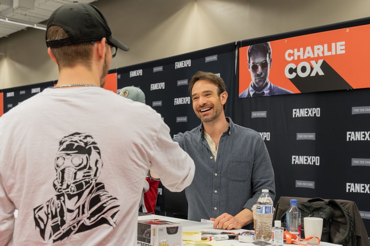 Charlie Cox, wearing a blue button-up shirt and sporting short brown hair and a trimmed beard, smiles warmly as he shakes hands with a fan across the autograph table. The fan, seen from behind, wears a black baseball cap, glasses, a silver chain, and a white long-sleeve shirt featuring a black illustration of Star-Lord on the back. The table holds water bottles, pens, and Funko Pop boxes. Behind Charlie is a bold red-and-black banner with his name and an image of him as Daredevil, set against a black FAN EXPO backdrop.