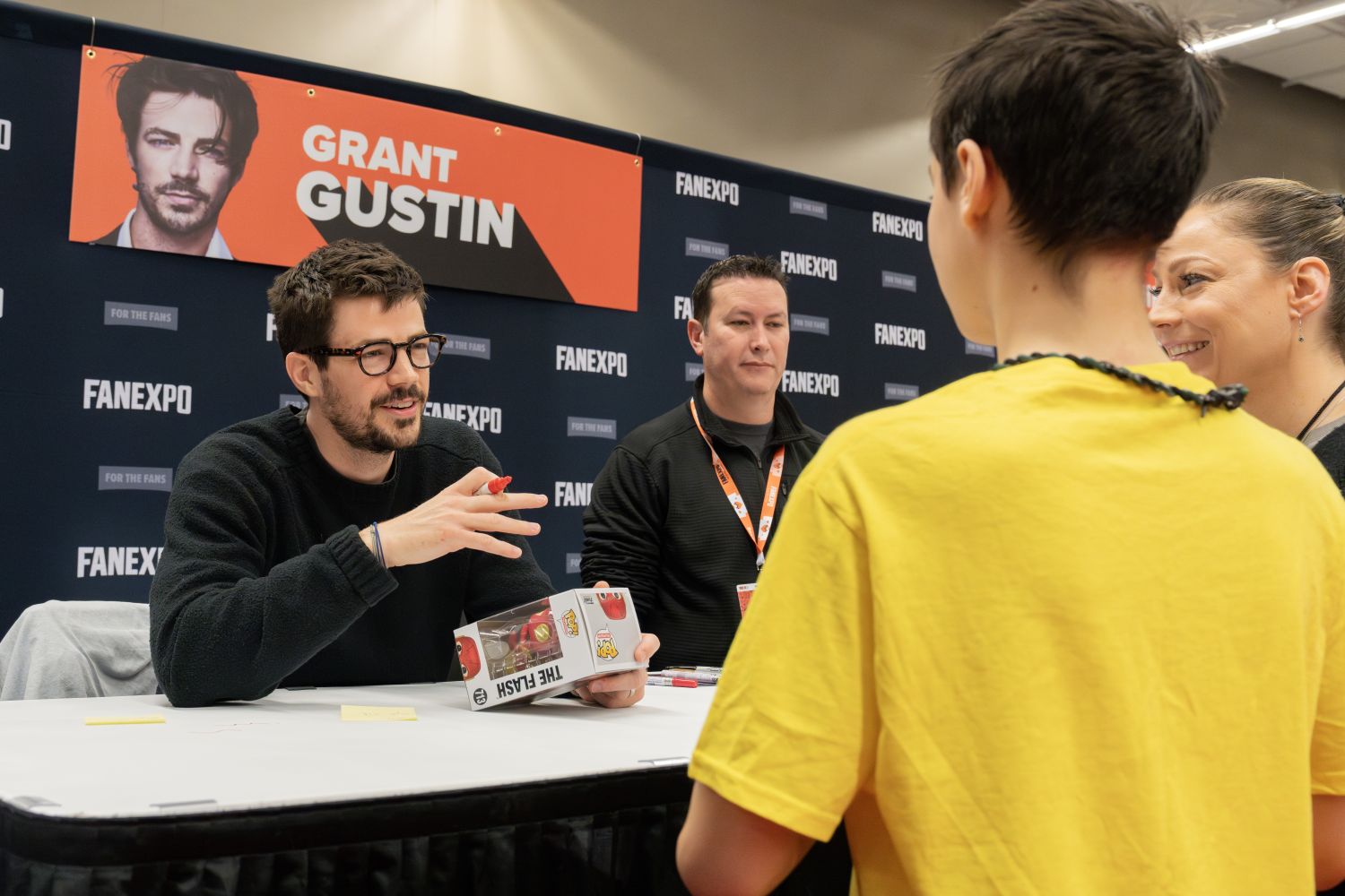 Grant Gustin, wearing black-framed glasses and a black sweater, gestures with a red Sharpie while smiling and talking to a young fan in a bright yellow shirt. He holds a Flash Funko Pop figure box. Grant's dark hair is slightly tousled, and he’s seated at a white table with another staff member in a black hoodie beside him. The FAN EXPO banner behind him features his name and photo with tousled hair and a thoughtful look.