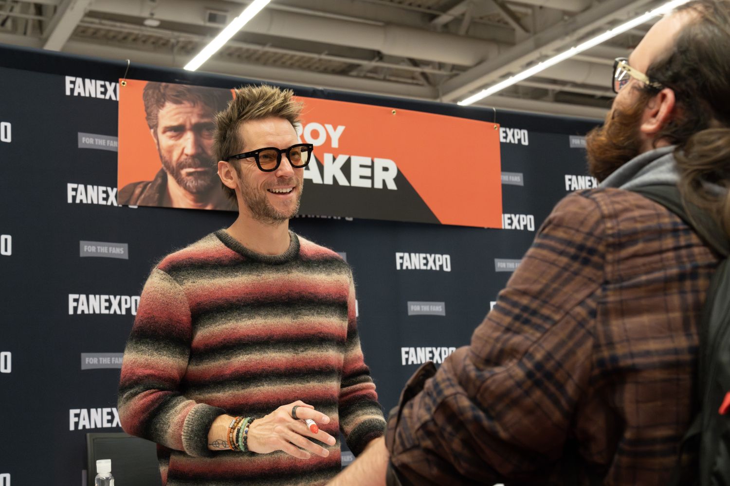 Troy Baker stands behind his autograph table wearing a multicolored striped sweater, dark-rimmed glasses, and bracelets on both wrists. He smiles at a fan in a brown plaid flannel shirt and glasses, who is standing across from him. Troy’s hair is styled up in a spiky fashion and his trimmed beard matches the rugged portrait of him on the banner above, which reads “Troy Baker” in bold white letters on orange and black. The FAN EXPO background features repeated logos.