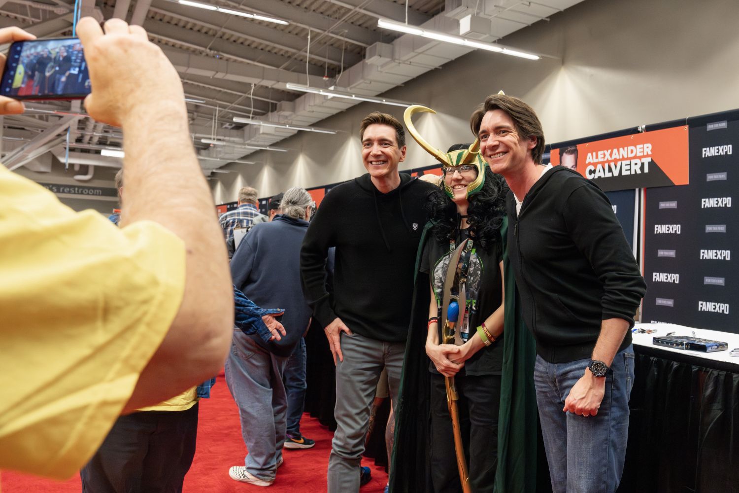 James and Oliver Phelps, both wearing black hoodies and jeans, smile while posing with a cosplayer dressed as Loki—complete with green and black robes, golden horns, and a staff. They stand close together on the red carpet of the FAN EXPO autograph area. Another attendee in a yellow shirt holds up a phone to snap a picture. Behind them is a banner for Alexander Calvert, and several fans wait in line along the back wall.
