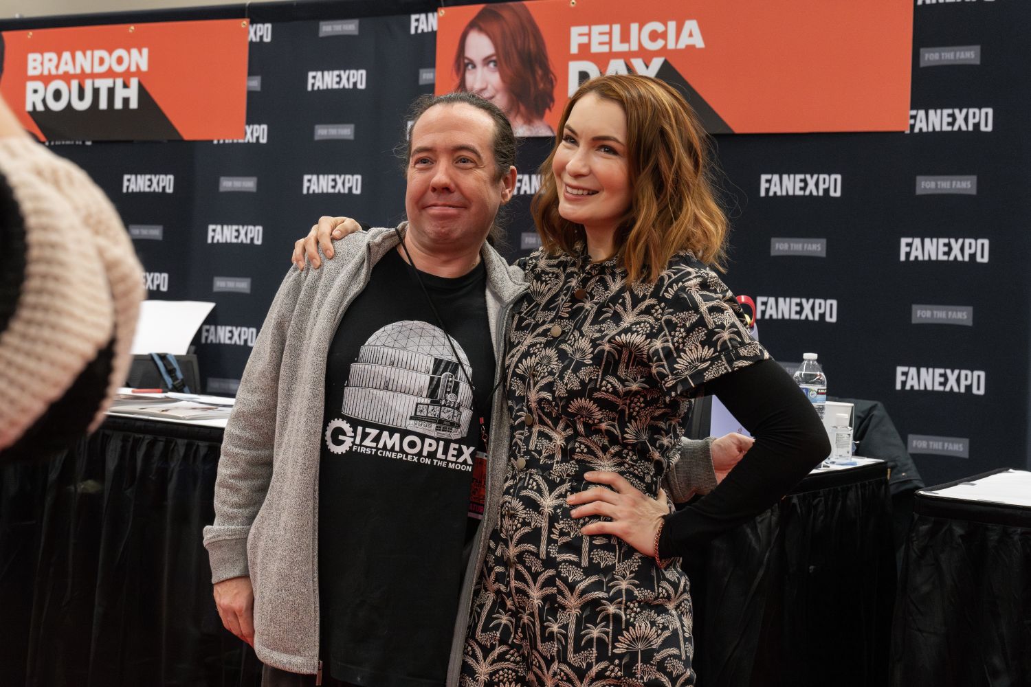 Felicia Day, wearing a black and white floral jumpsuit with short sleeves and auburn hair styled in loose waves, smiles with her arm around a fan. The fan wears a gray zip-up hoodie and a black shirt with a ‘Gizmoplex’ graphic. They stand in front of Felicia’s table, which is marked with her name and a photo of her smiling brightly. Brandon Routh’s nearby banner is also visible in the background, along with FAN EXPO signage.