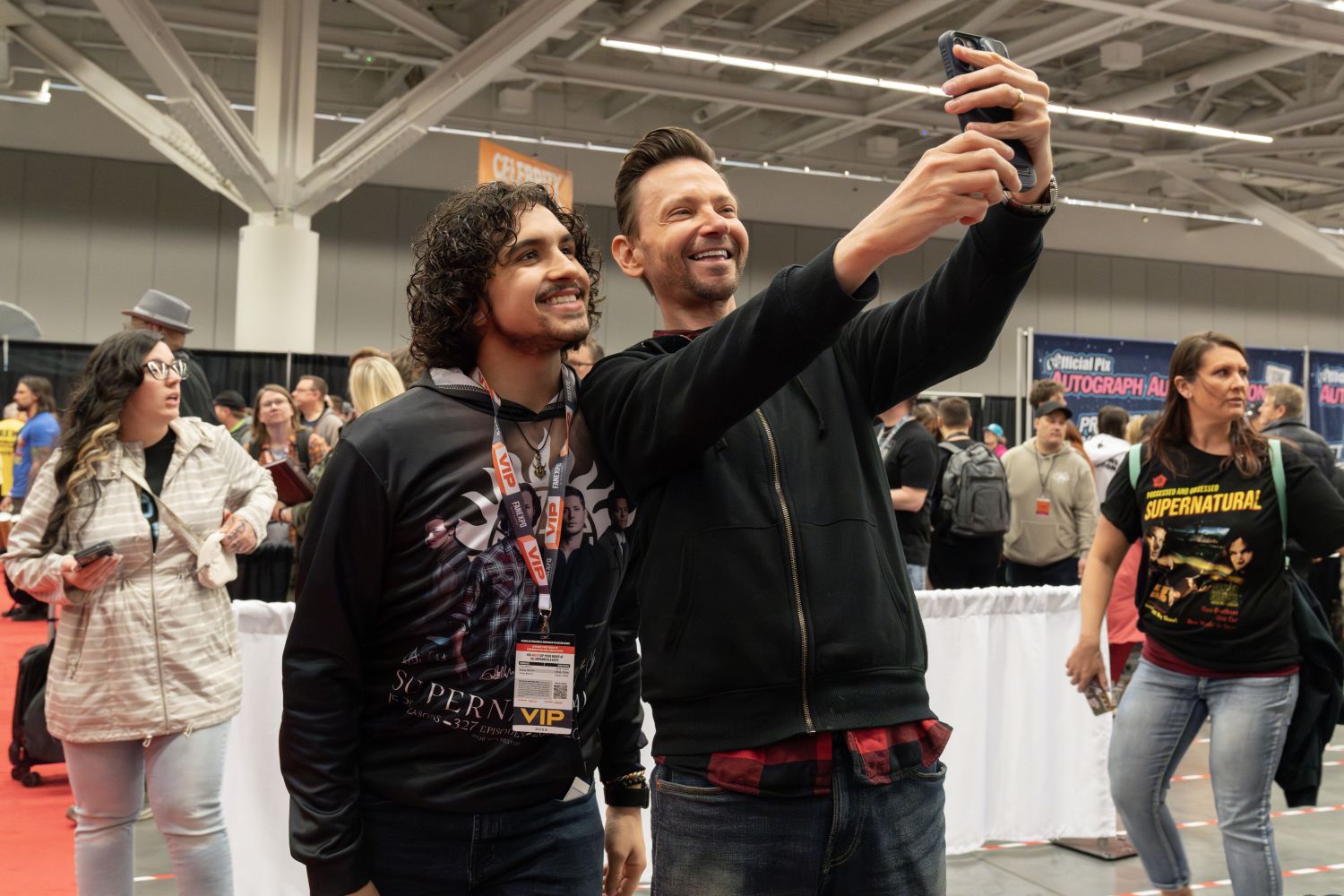 An excited fan in a black Supernatural hoodie with curly dark hair poses for a selfie with DJ Qualls wearing a black hoodie and red flannel shirt. The actor holds the phone high and grins as they both smile for the shot. The FAN EXPO show floor is bustling behind them, with other fans walking and chatting. A woman in a Supernatural shirt walks by holding her phone, while others in the background wait in lines near the Celebrity Autograph area.