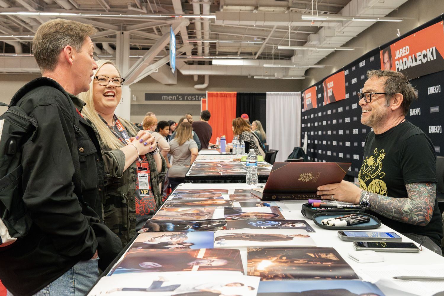 Mark Sheppard, in a black t-shirt with gold lettering and colorful sleeve tattoos, laughs while speaking to a couple at his autograph table. The couple wears FAN EXPO VIP badges; the woman has platinum blonde hair and purple nails, wearing camouflage and glasses, while the man wears a black jacket. The table is filled with glossy 8x10 photos from Supernatural. Behind Mark is a banner with Jared Padalecki’s name, and other fans line up along the autograph area.