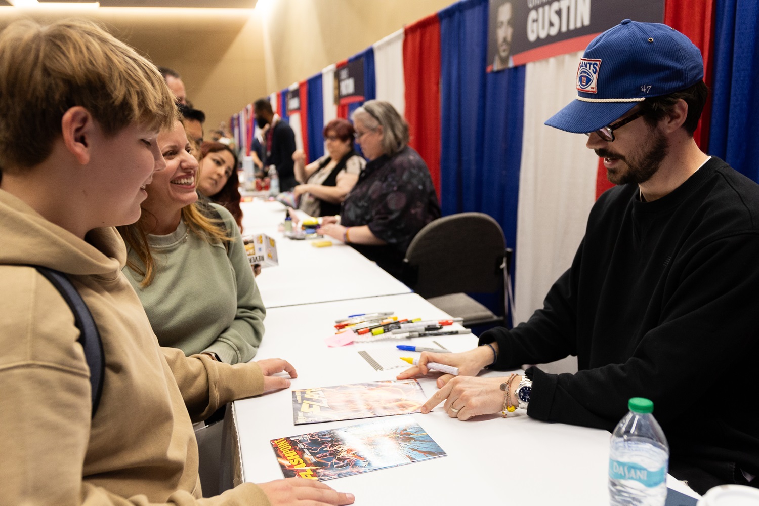 Grant Gustin signs an autograph for two fans at his table at a FAN EXPO event, both of whom are smiling. Various markers and pens are scattered on the table.
