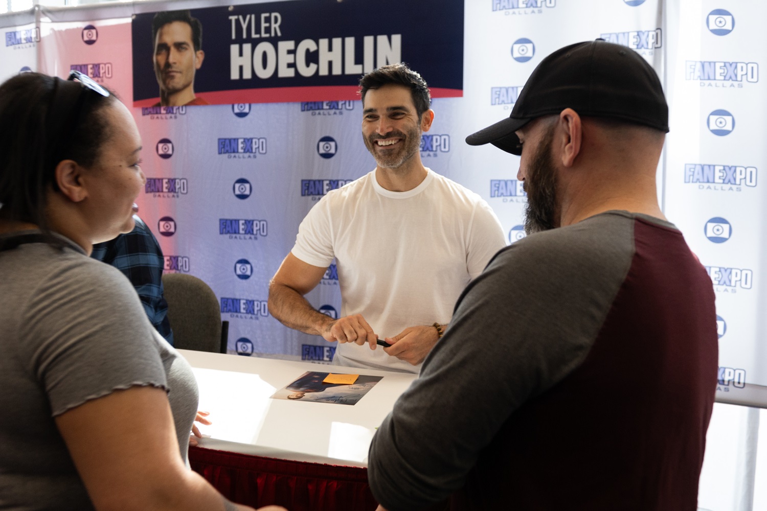 Tyler Hoechlin, smiling and wearing a white t-shirt, engages with fans at his autograph station at a FAN EXPO event, with his name banner visible in the background.