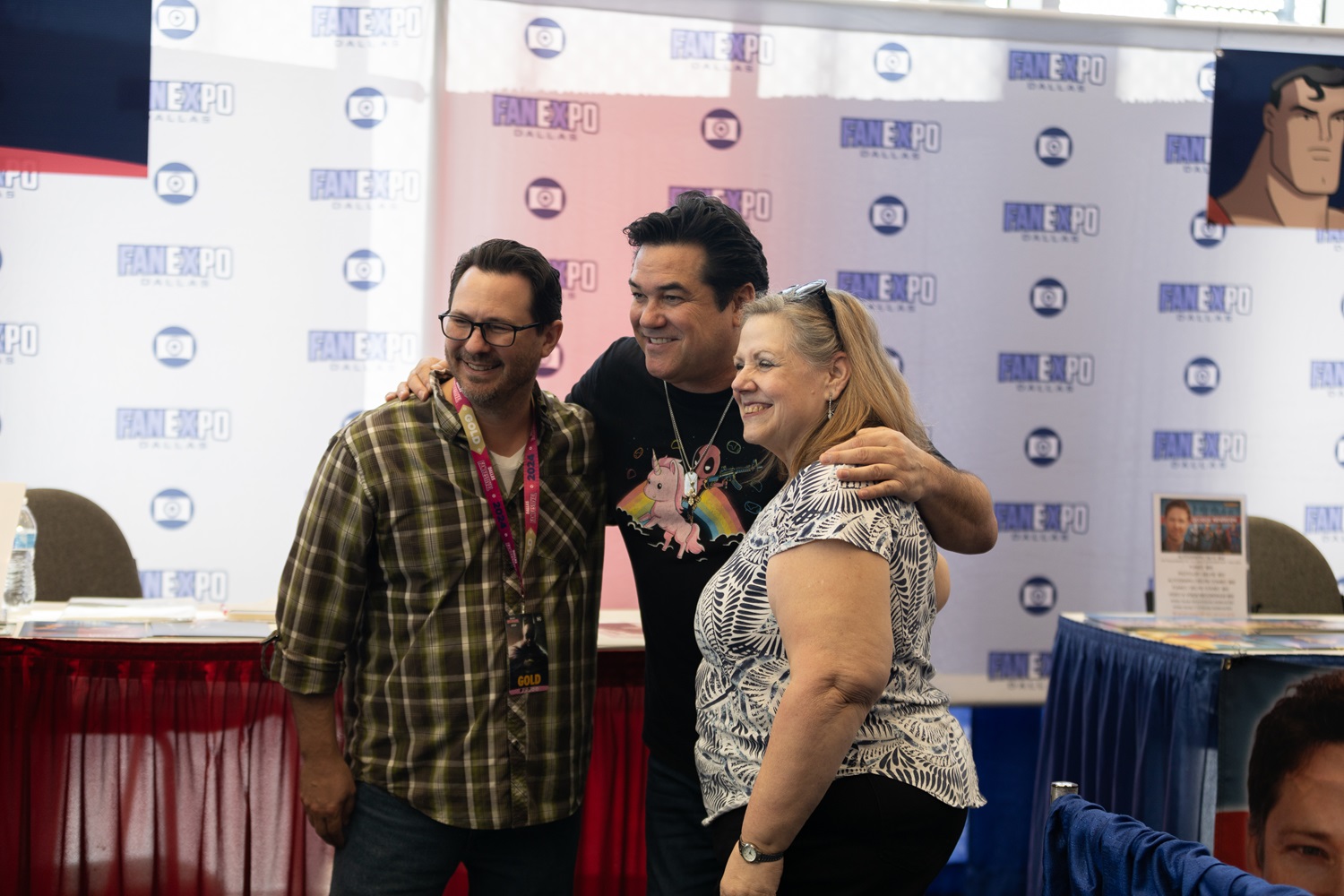 Dean Cain, smiling and wearing a colorful unicorn t-shirt, poses with two fans at Dallas FAN FESTIVAL in front of his autograph station.