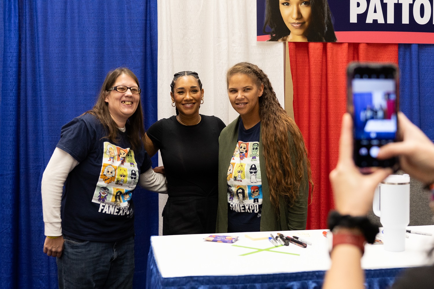 Candice Patton, smiling, poses with two fans at her table at a FAN EXPO event, while another person takes a photo on their smartphone.