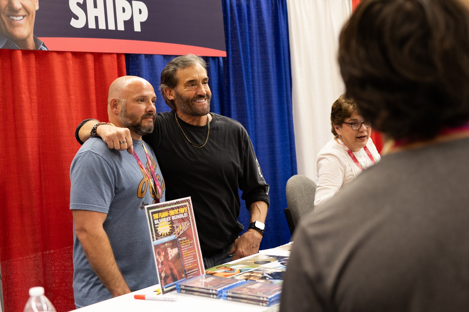 John Wesley Shipp poses with a fan at his table at a FAN EXPO event. Both are smiling, and an event assistant sits nearby.