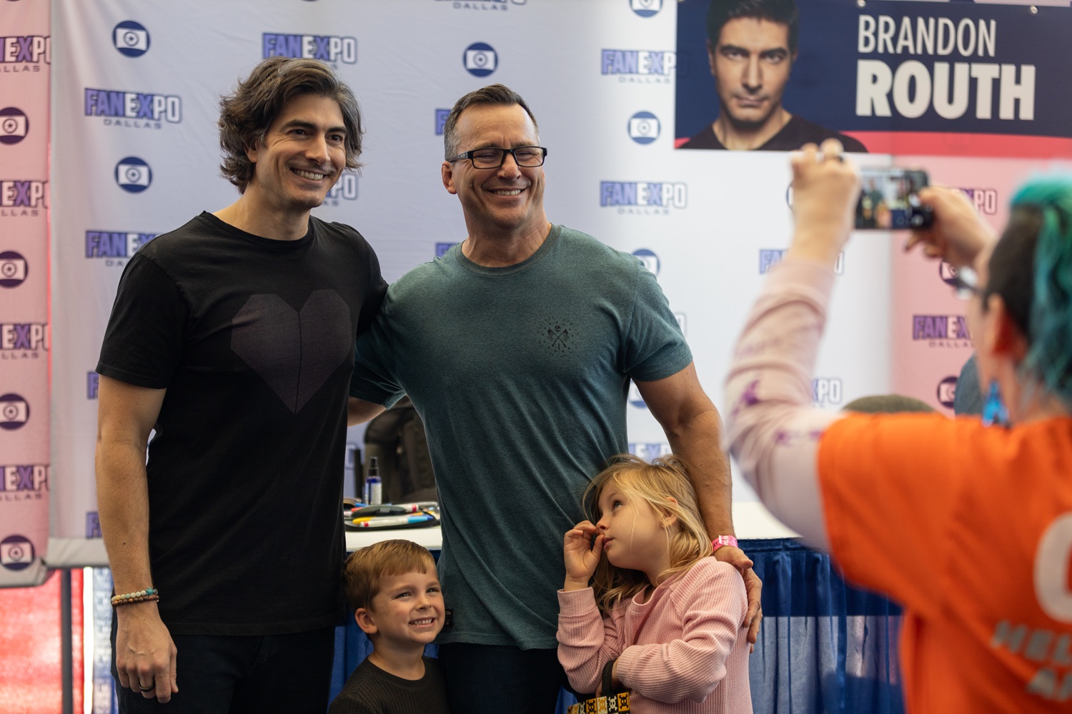 Brandon Routh, smiling widely, poses with an adult fan and two young children in front of his autograph station at Dallas FAN FESTIVAL.