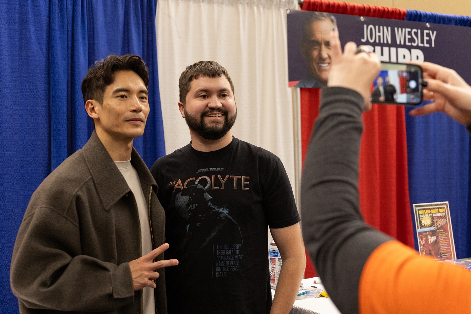 Manny Jacinto, dressed casually, poses with a fan, holding up a peace sign at his table at a FAN EXPO event.