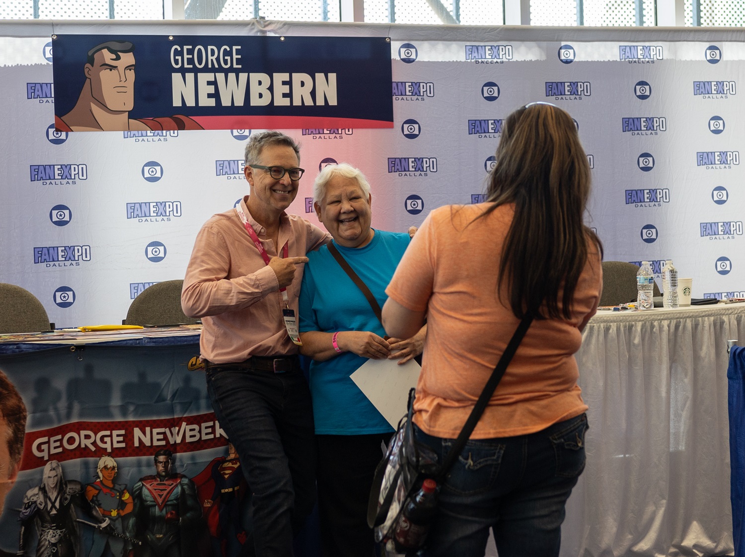 George Newbern, smiling and pointing at a fan, poses for a photo at his booth at Dallas FAN FESTIVAL, with a banner featuring his name and a Superman illustration above.