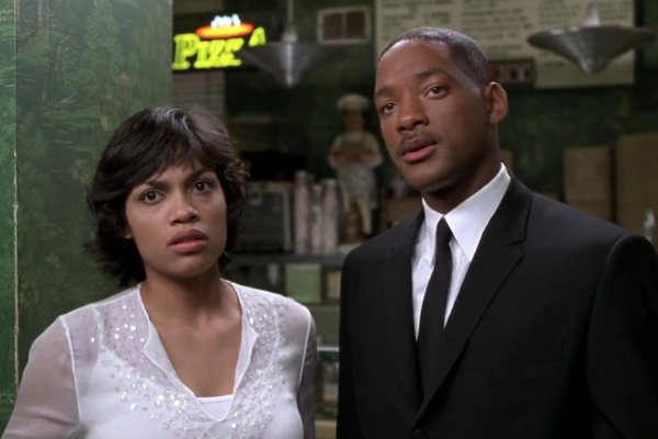 Rosario Dawson in a white blouse standing next to Will Smith in a suit, both looking exasperated/"Are you serious" face. Behing them is a stall for a pizza place.