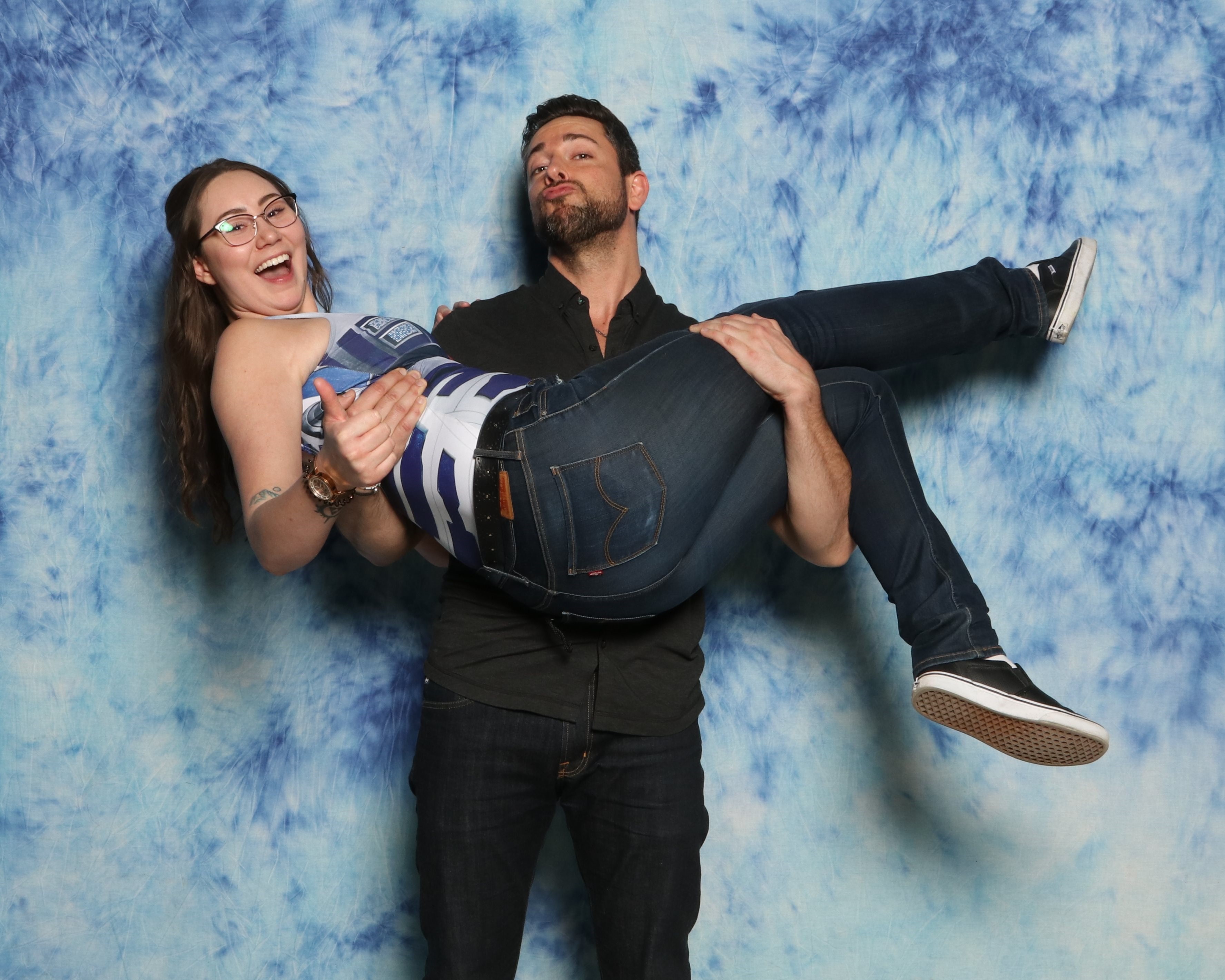 Zachary Levi stands against a blue background and holds up Karli, a brunette that is wearing a blue and white tank top with blue jeans and black and white sneakers. Zachary is wearing blue jeans and a black dress shirt.