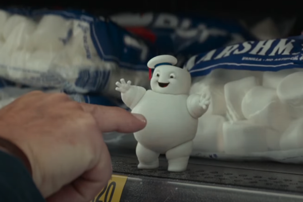 A mini Staypuft Marshmallow man in a cute sailor hat, is smiling as it gets poked in the belly on the shelf of a grocery store.