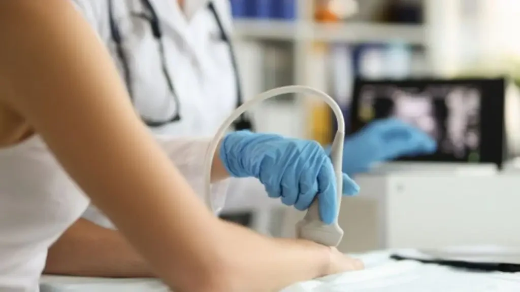 Médica com luvas azuis realiza exame de ultrassom no braço de uma paciente para diagnóstico precoce de doenças imunomediadas. Ao fundo, tela mostra o resultado da imagem médica.