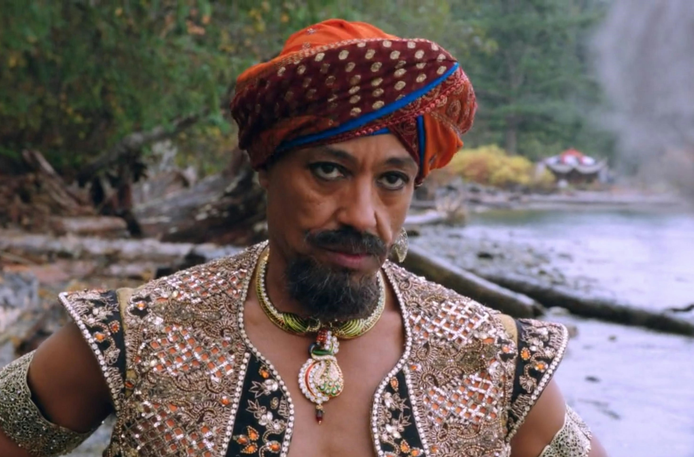 Giancarlo as a genie. He is standing by the side of a river in a red turban, ornate vest, and wearing a large necklace and earring.