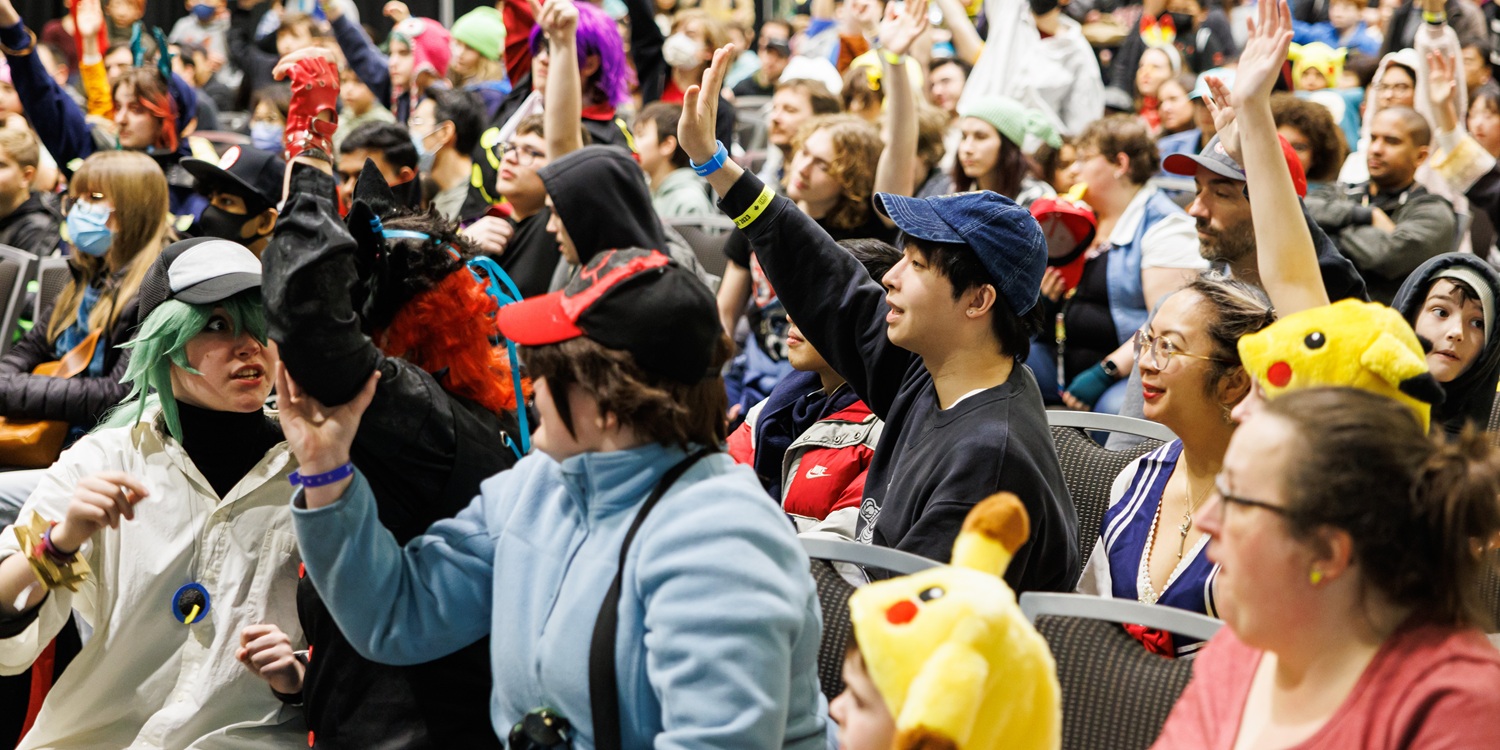 A crowd of attendees gathers closely together, raising hands and cheering during a trivia event at FAN EXPO Cleveland. Fans wear casual clothing and cosplay as they participate in an interactive game.