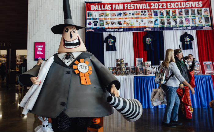 A large figure of the Mayor character from "The Nightmare Before Christmas" stands prominently in a convention hall. The Mayor holds a black and white striped megaphone and wears his signature top hat, suit, and a badge labeled "MAYOR". In the background, attendees browse a booth with "DALLAS FAN FESTIVAL 2023 EXCLUSIVES" and various merchandise on display. A sign reading "EXHIBIT HALL" is seen to the left.