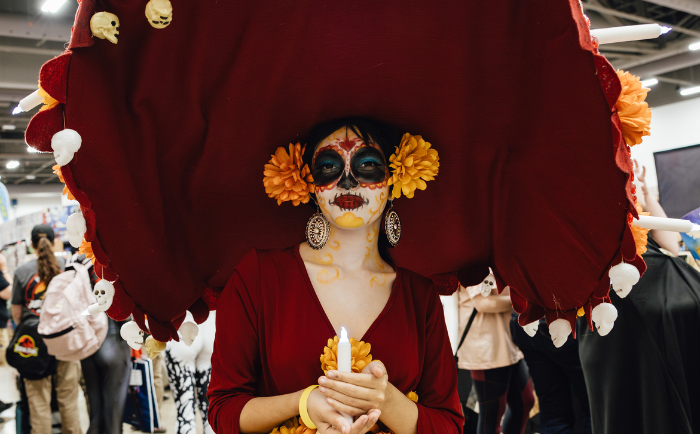 A woman dressed in a Day of the Dead inspired costume stands in a convention setting. She wears vibrant makeup resembling a sugar skull, with red, black, and yellow hues. Floral decorations adorn her face and golden earrings dangle from her ears. She holds a yellow candle in her hands, and her attire includes a large deep red hat adorned with small skulls and orange flowers. The bustling background shows other convention-goers and booths.