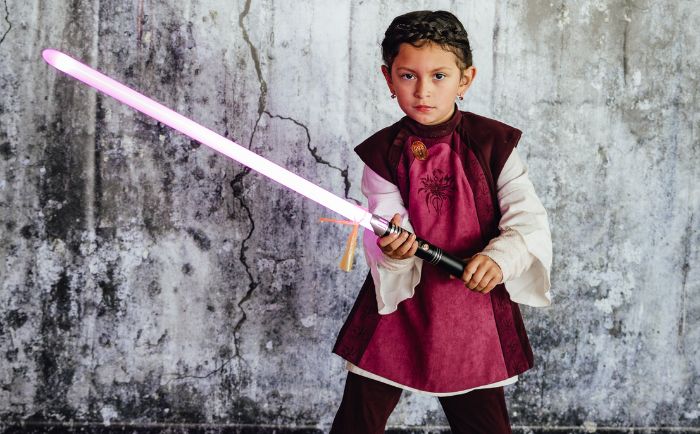 A young girl confidently poses in front of a weathered gray wall. She is dressed in a maroon vest with intricate designs, a white long-sleeve shirt underneath, and matching maroon pants. She holds a bright pink glowing lightsaber in her right hand, her expression intense and focused. The lightsaber casts a soft pink hue onto her determined face.