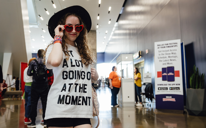 A young woman with curly hair stands in a modern indoor setting, adjusting heart-shaped red sunglasses. She dons a black hat and a white T-shirt with the text "GOT A LOT GOING ON AT THE MOMENT" printed in bold. She has various colorful bracelets on her wrist. In the background, attendees can be seen walking, and there is an information sign for the "Dallas Fan Festival" to the right. The atmosphere is lively and bustling.