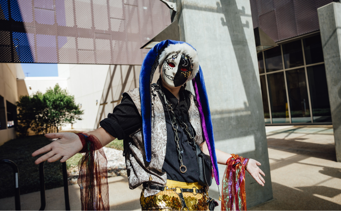 A person in a vibrant costume stands outdoors in a courtyard, posing with arms outstretched. They wear a mask with a painted face, reminiscent of a skull, and a blue hood lined with fur. Their attire consists of a patterned shirt, shiny gold pants, multiple necklaces, and colorful streamers attached to their wrists. The surrounding area features modern architecture, including concrete pillars and a mesh-like facade, contrasting with the individual's eclectic appearance.
