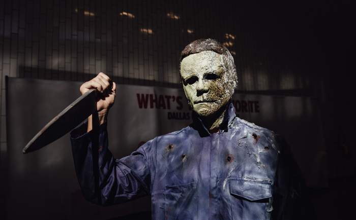 A figure wearing a weathered mask and blue jumpsuit is dressed as Michael Meyers and stands menacingly with a raised knife. The backdrop includes a wall and a sign reading "WHAT'S YOUR FAVORITE FAN FEST MEMORY", suggesting an event setting. The ominous lighting casts shadows, enhancing the eerie atmosphere