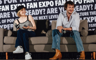 Grace Van Dien (left) and Joseph Quinn (right) sitting on a couch, smiling and holding microphones at a main stage panel. Grace is wearing a black hat, a leopard print tank top, blue jeans, and white sneakers. Joseph Quinn is wearing a light blue, button down shirt, blue jeans, and brown cowboy boots.