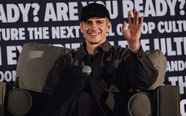 Hayden Christensen sitting on a grey armchair, smiling and waving at the audience. He is wearing a black baseball cap, and a black button down shirt.