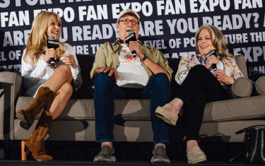 Christie Brinkley (left), Chevy Chase (middle), and Beverly D'Angelo (right) sitting together on a couch laughing and smiling during a main stage panel. Christie is wearing a white dress with brown cowboy boots, Chevy is wearing a white baseball cap, white t-shirt with a beige blazer, blue jeans, and sneakers. Beverly is wearing a floral blouse, black jeans, and beige shoes.