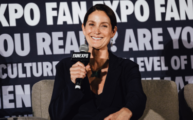 Carrie-Anne Moss sitting on stage, smiling with a microphone in hand. She is wearing a black blazer with a black blouse, and earrings.