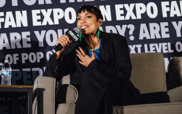 Rosario Dawson sitting on a couch on stage, looking off to the left while talking into a microphone. She is wearing green earrings, a blue blouse, and a black suit.