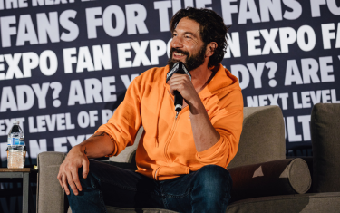 Jon Bernthal sitting on stage, laughing into a microphone during a panel. He is wearing an orange hoodie and blue jeans.