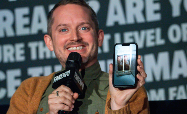 The image shows Elijah Wood at a FAN EXPO panel, enthusiastically speaking into a microphone and holding up a smartphone displaying a photo of two candles. He has a joyful expression and is dressed in a casual button-up cardigan over a green shirt. The background includes a sign with FAN EXPO branding, indicating the event's focus on fan interaction. His engagement with the audience and his photo display suggest a personal or interactive segment of the panel discussion.