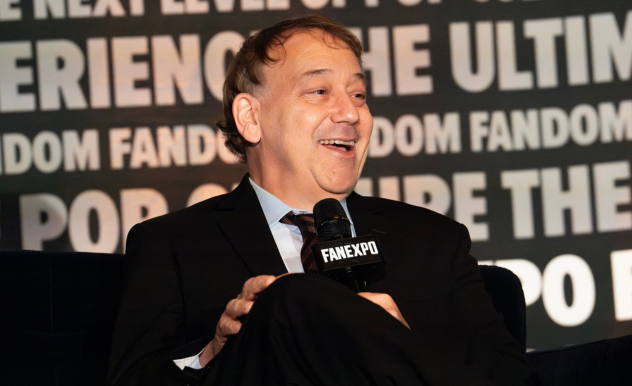 The image captures Sam Raimi speaking at a FAN EXPO panel, clearly enjoying the moment. He is dressed in a formal black suit and tie, holding a microphone, and smiling broadly as he engages with the audience. The backdrop features phrases like "ULTIMATE FANDOM EXPERIENCE" and "POP CULTURE," emphasizing the event's theme. His relaxed and joyful demeanor suggests he is discussing something amusing or entertaining, contributing to the lively atmosphere of the panel.