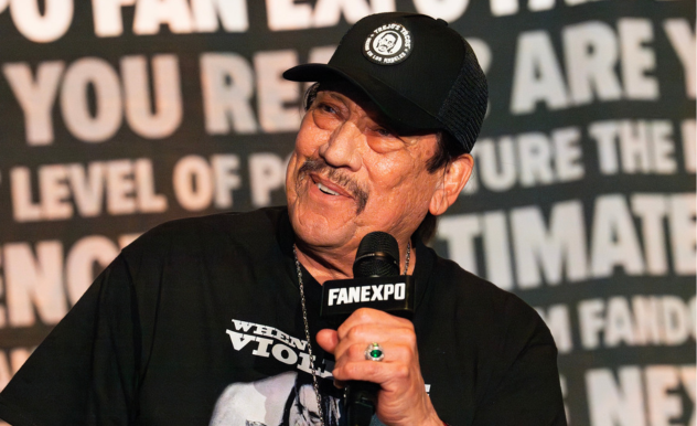 The image features Danny Trejo speaking at a FAN EXPO panel, displaying a warm and engaging smile. He is dressed in a casual black t-shirt with a graphic design and a black cap with a logo, holding a microphone. His joyful expression and animated gestures suggest he is sharing an amusing story or interacting positively with the audience. The backdrop includes bold text emphasizing the fan-centric nature of the event, enhancing the lively and inclusive atmosphere of the panel.