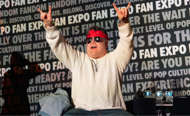 The image captures Sean Astin enthusiastically raising his arms in a celebratory gesture at a FAN EXPO panel. He is wearing a cream-colored cable-knit sweater and jeans, accessorized with a red bandana over his eyes, which adds a playful and energetic element to his appearance. His open-mouthed smile and animated pose suggest he is either reacting to a cheering crowd or participating in an exciting panel discussion. The backdrop is filled with text related to fandom and pop culture, emphasizing the vibrant and interactive nature of the event.