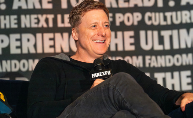 The image features Alan Tudyk speaking at a FAN EXPO panel, displaying a relaxed and amused expression. He is dressed casually in a black sweater and dark pants, sitting comfortably with one leg crossed over the other. He holds a microphone, suggesting he is either responding to a question or sharing an anecdote, contributing to the interactive and engaging atmosphere of the event. The backdrop, filled with texts about fan experiences and pop culture, reinforces the theme of the panel discussion.