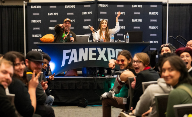 The image depicts a lively and interactive panel at FAN EXPO. Two panelists, a man and Sarah Natochenny, are seated at a table on stage, smiling and engaging with the audience. The man, wearing glasses and a green hat, waves a plush toy while Sarah, with long hair, gestures excitedly. The audience in the foreground appears animated and happy, with some pointing and making thumbs-up gestures towards the camera. The setting is casual, emphasizing the friendly and engaging nature of the panel discussions at the event. The backdrop prominently displays the FAN EXPO logo, underscoring the event's branding and theme.