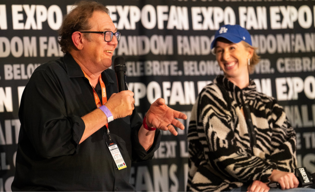 The image features Fred Tatasciore and Maggie Robertson participating in a panel at FAN EXPO. Fred, speaking into a microphone, is gesturing with his hand as if explaining a point or sharing a story, wearing a black shirt and glasses. Beside him, Maggie, dressed in a zebra-striped sweater and a blue baseball cap, listens with a delighted smile, clearly enjoying the conversation. The backdrop is filled with text promoting the event’s themes of fandom and pop culture, setting the scene for a dynamic and engaging discussion.