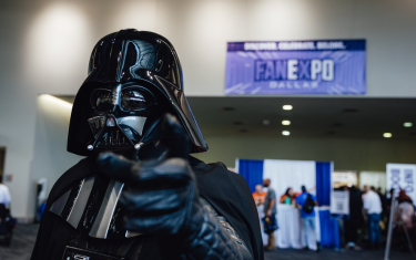 a cosplayer dressed as Darth Vader looking and pointing at the camera, with a blurred FAN EXPO Dallas banner in the background.