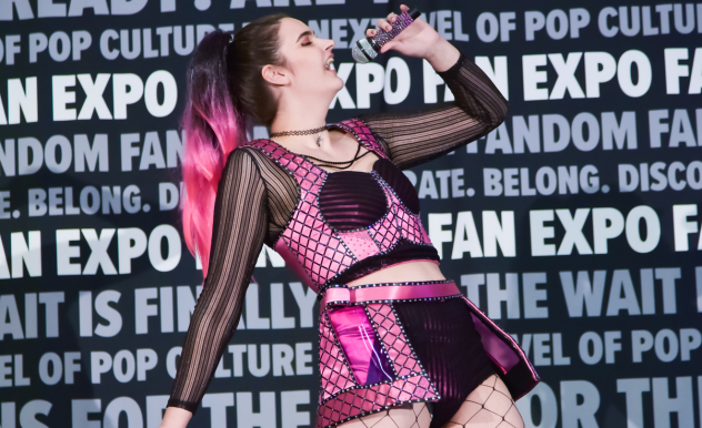 The image depicts a vibrant and energetic moment at FAN EXPO, featuring a woman performing onstage. She is dressed in a futuristic-looking costume consisting of a black and purple fishnet outfit with shiny pink accents and a stylish skirt. Her hair is styled in a long ponytail with pink highlights, enhancing her dynamic look. She is singing into a microphone, her head tilted back in a pose that captures her passion and energy, making the performance feel lively and engaging. The background is filled with text promoting the expo, which highlights the event's celebration of pop culture and creativity.