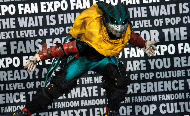 The image captures a dynamic and intense moment at FAN EXPO, featuring a person in a highly detailed and dramatic costume. The costume includes a menacing dinosaur-like mask with sharp teeth and a vibrant color palette of teal, red, and yellow. The cosplayer is posed in a crouching position, with arms outstretched and clawed hands, adding to the fierce and theatrical nature of the costume. The backdrop is filled with text related to the expo, which emphasizes the celebration of fandom and pop culture. This setting provides a perfect stage for such an elaborate and creatively inspired cosplay display.