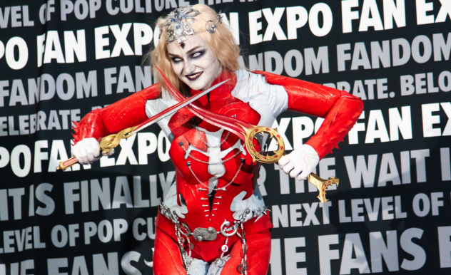 The image showcases a person in an elaborate and striking cosplay at FAN EXPO. The costume is vividly detailed, featuring a red and white bodysuit with intricate designs and attachments, including chains and ornamental keys. The cosplayer is styled with dramatic makeup and a complex headpiece, completing the fantastical character look. They are holding a violin bow and a violin with a creative, ornate design, adding a unique and theatrical element to the ensemble. The backdrop filled with event-related text emphasizes the vibrant and creative atmosphere of the expo, highlighting the artistry and passion of the cosplay community.