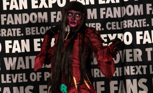 The image features a person in an intense and eerie cosplay at FAN EXPO. The costume includes a dark, tattered robe and long, stringy black hair, creating a haunting appearance. The cosplayer's face is painted in deep red and black, enhancing the sinister aspect of the character. They are standing with arms outstretched and fingers splayed, adding to the dramatic and spooky vibe of the portrayal. The backdrop, filled with expo-related text, sets a contrasting scene that highlights the imaginative and diverse expressions of cosplay within the fan culture celebrated at the event.