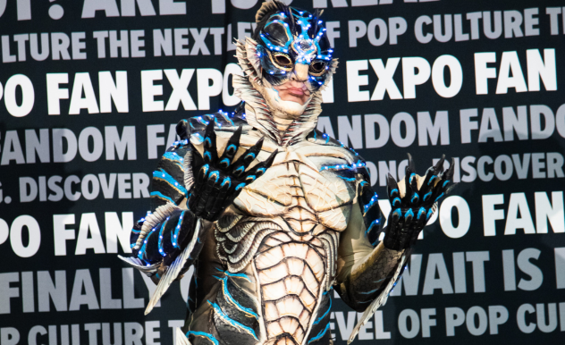 The image features an exceptionally detailed and intricate cosplay at FAN EXPO. The cosplayer is dressed in a creature-like suit that resembles an armored sea creature, with scales and fins, predominantly in tones of white, black, and striking blue highlights. The costume includes elaborate facial makeup and sculpted pieces that cover the eyes, adding a layer of mystery and otherworldly appeal. The cosplayer's hands are adorned with claw-like extensions, emphasizing the meticulous craftsmanship of the costume. This portrayal, set against a backdrop filled with event-related text, highlights the high level of creativity and artistry found in the cosplay community at such fan conventions.
