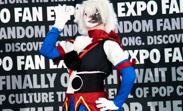 The image features a cosplayer at FAN EXPO dressed in a vibrant and detailed anime character costume. The outfit consists of a mix of bold colors, including red, blue, and black, with white fur accents around the collar and sleeves. The cosplayer's face is partially covered with a white and black mask, styled to mimic an animalistic look, and they wear a white wig that adds to the character's dramatic flair. They are striking a confident pose with one hand raised, showcasing their character's dynamic personality. The backdrop, filled with text related to the expo, emphasizes the exciting and creative atmosphere of the event, celebrating fandom and pop culture.