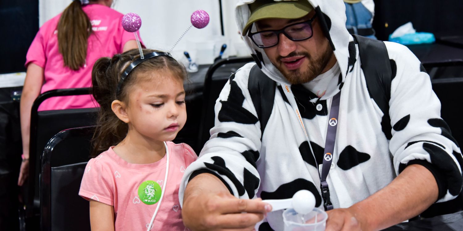 A young girl in a pink shirt with sparkly antenna headband watches attentively as a man dressed in a cow-print onesie and glasses carefully measures liquid into a small cup. They are seated at a table in a workshop setting at the convention.