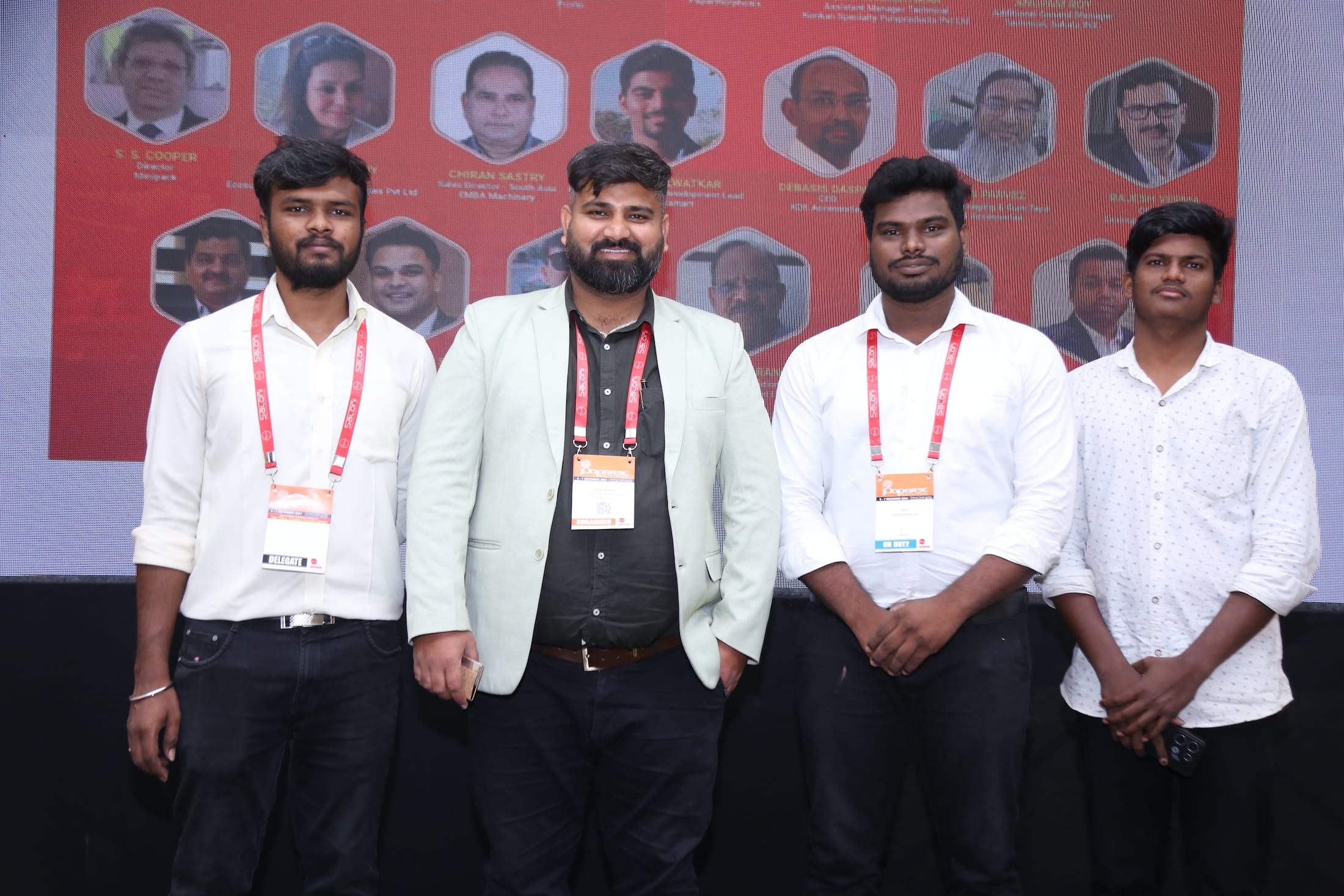 Four men wearing lanyards posing in front of a red banner featuring speaker headshots at Paperex South India.