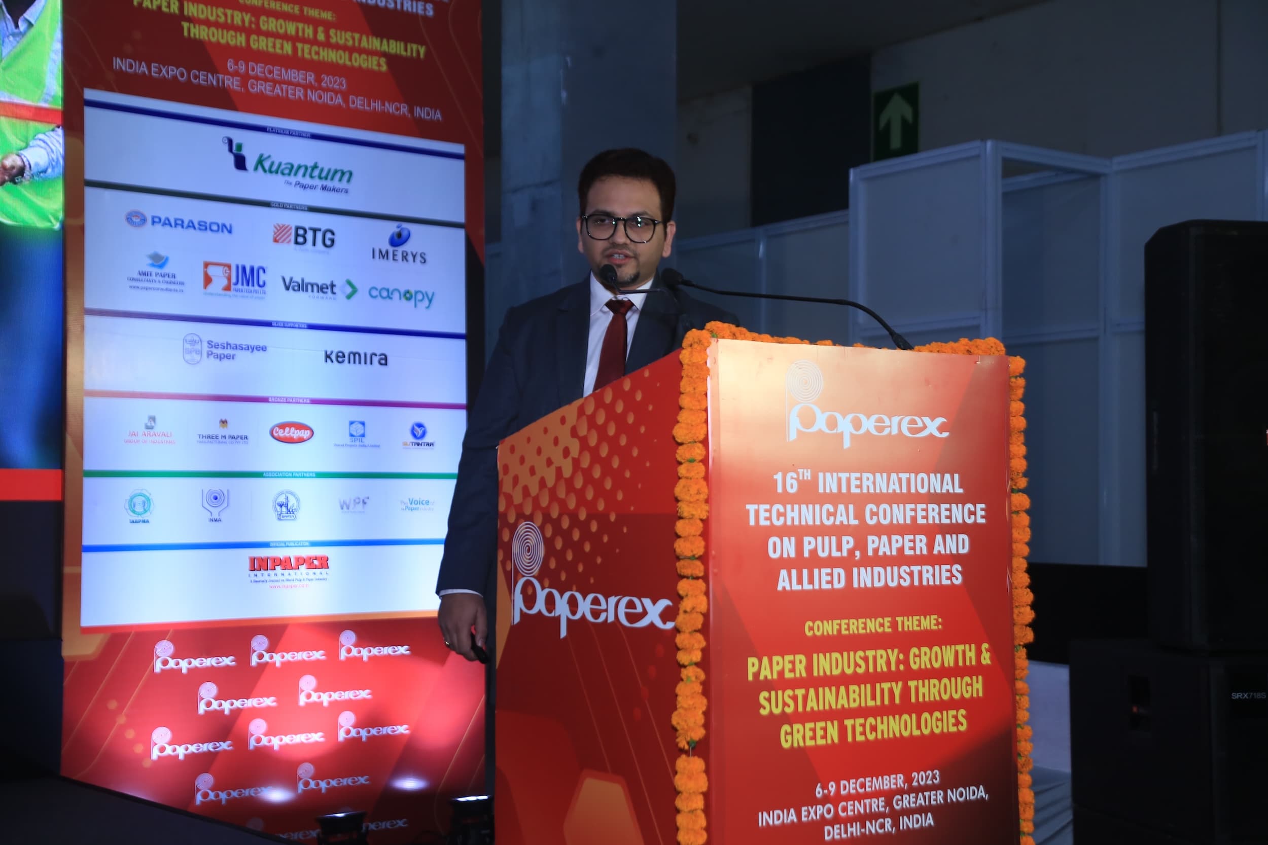 Man speaking at a paper industry conference podium with sponsor banners displayed